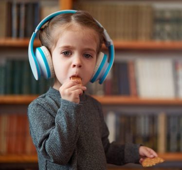 Small kid in a grey shirt, eating an oval biscuit. They are wearing white and cyan headphones.