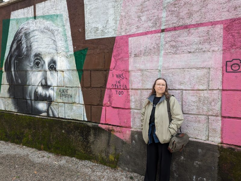 Woman in a heavy jacket leaning against a pink painted wall next to a mural of Albert Einstein.