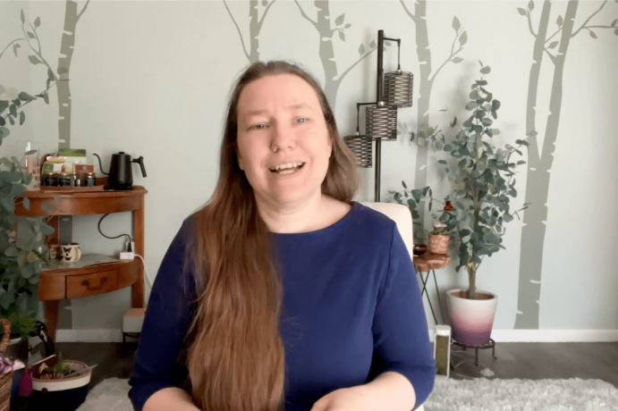 Heather in office A pale skinned woman in a dark blue shirt with long hair, talking at the camera and smiling. Behind her is a pale green wall with painted trees in darker green.