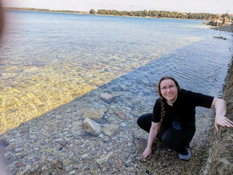 PXL_20240406_093058457 A person with braided brown hair smiling at the camera while crouching on rocks next to a large expanse of water.