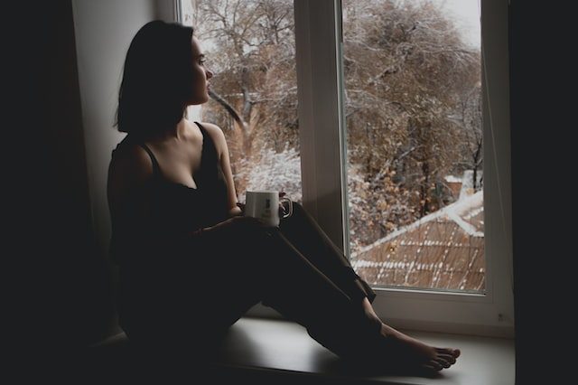Person covered in shadow, sitting on a window sill. They are looking out the window.