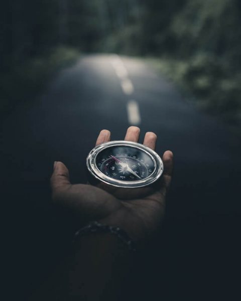 Close up on a hand holding out a silver compass, with a blurred out road in the background.