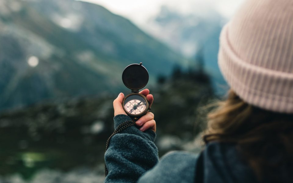 Someone holding up their hand with a compass in it, pointed towards the blurred mountains in the background.