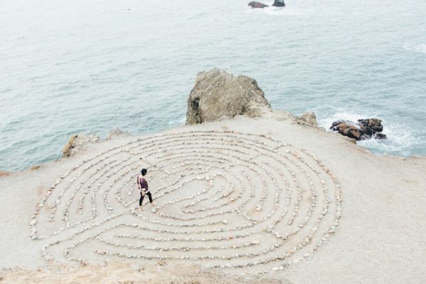 A circular maze in the sand made out of rocks. It is near water and there is a person in a red shirt walking through it.