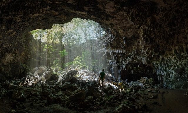 Giant cave with stalactites and algae. There's light beaming through a big hole in its roof and a person, in the distance, looking up.
