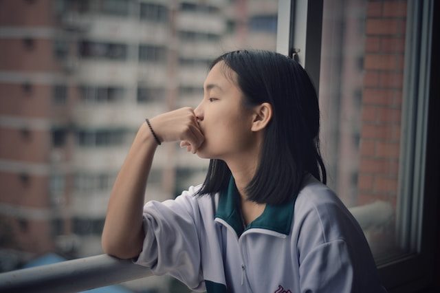 Asian female presenting person wearing a sweater sitting at a window and looking out with unfocused gaze and head leaning on her hand as if thinking.