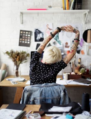 A person stretching their arms above them as they sit in a desk chair. The room around them is full of paper and drawings.