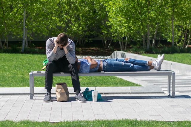 Two people on a bench with bushes behind them. One person is laying down with their head up against the other person, who is sitting and looking into a brown bag.