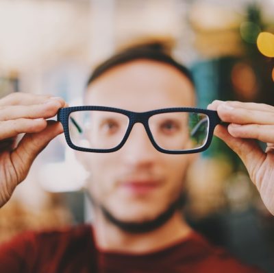 Person holding a pair of glasses out in front of their face, and the glasses are in focus and the person and background are blurry.