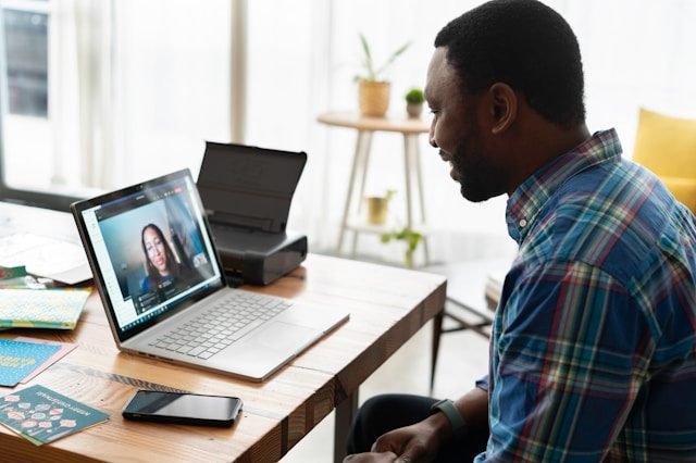 A person smiling and facing towards a laptop rested on top of a wooden table. The laptop displays a video call with another person.