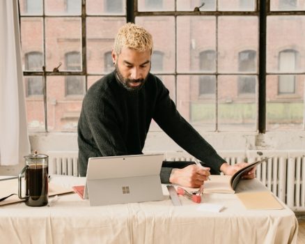 Someone seated at a white clothed table, looking down at their computer while writing down things in an open notebook.