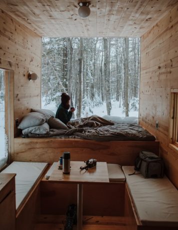 nachelle-nocom-51adhgg5KkE-unsplash Wide-shot of a person huddled up in a cabin with a wall opened up to the snowy trees.