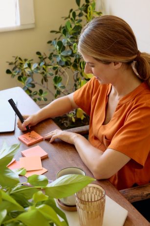 natalia-blauth-0qWmQ0M0v8M-unsplash A person sitting at a desk and writing on an orange sticky note.