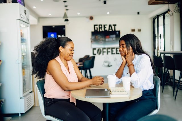 Two people sitting across from each other at a table, one of them is holding their hands up to the sides of their head and the other is resting their hands on the table.