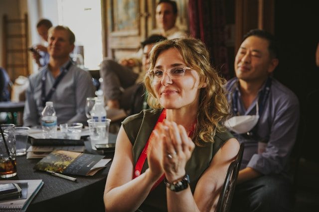 A woman with a lanyard sitting around a table with a couple other people. She is faced towards the camera and is clapping her hands.
