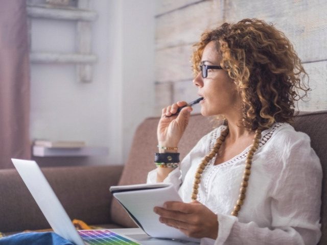 Person with brown curly hair, sitting on a couch. Holding a notepad in one hand, and holding a pen to their mouth in another.