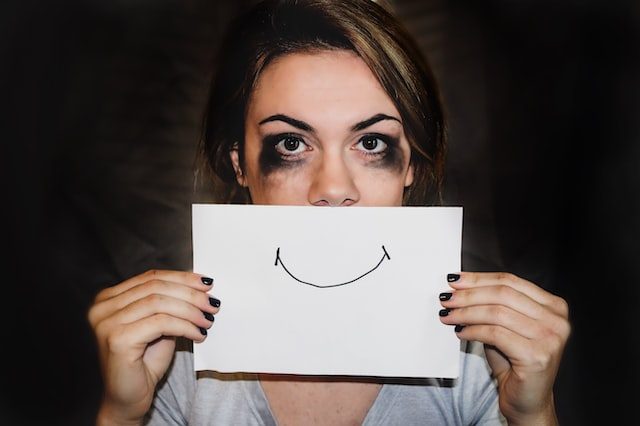 Woman with pale skin and dark shadows beneath her eyes holding up a drawing of a smile in front of her mouth.