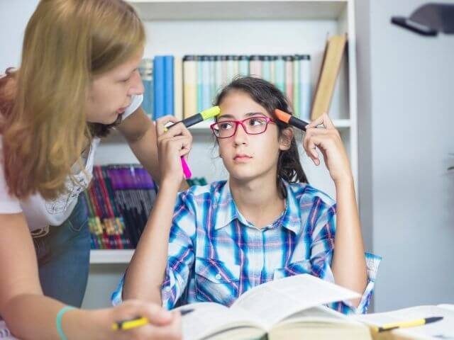 Person sitting at a desk with an open book. They are putting two pencils to their head and looking at the person in front of them.